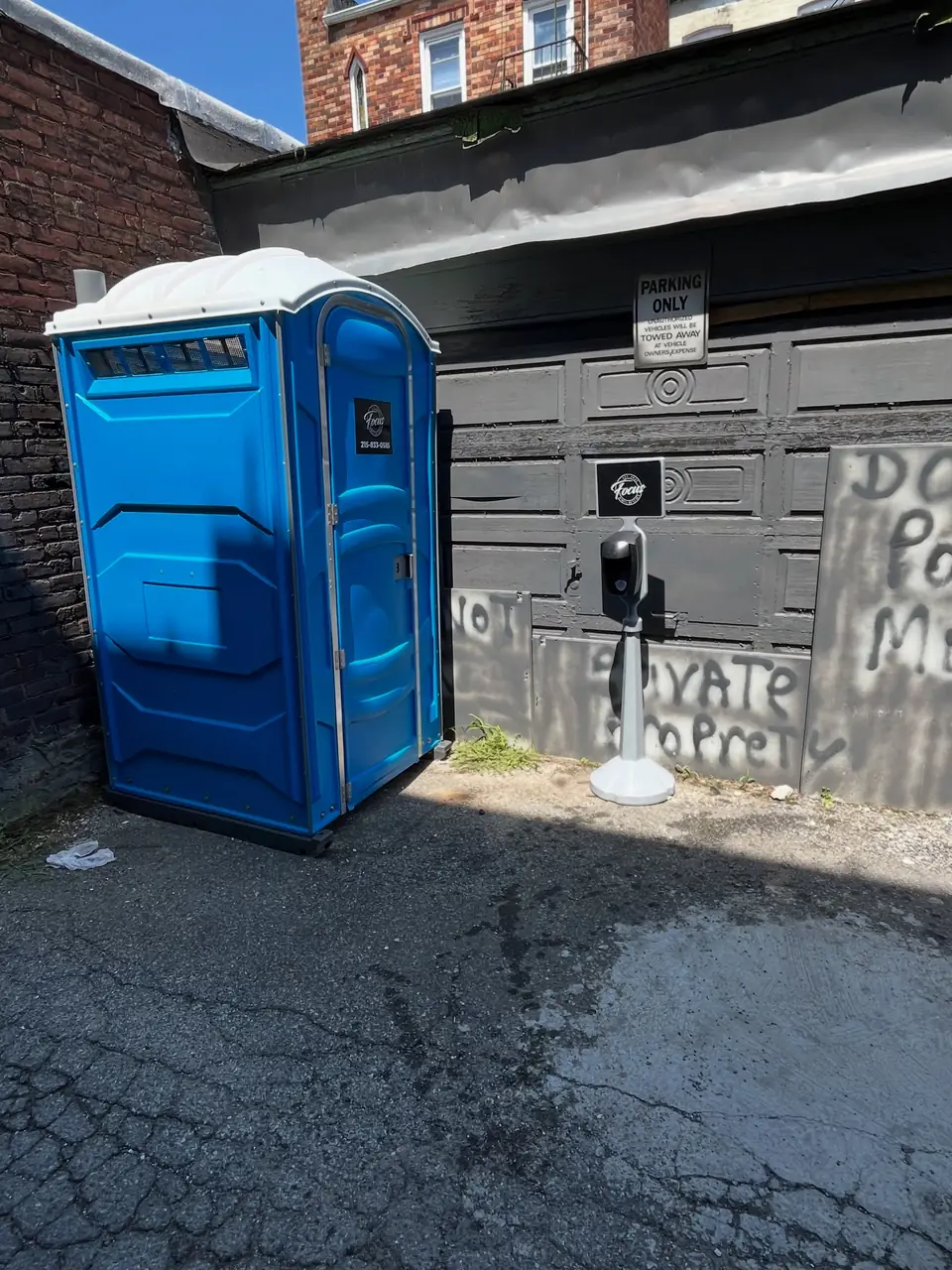 Blue standard porta potty placed beside a building on a paved alley