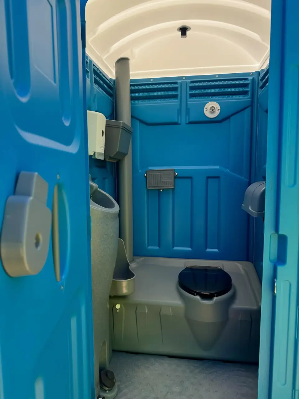 Inside a blue standard porta potty showing the vent pipe, urinal and toilet seat